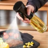 Hand pressing an oil sprayer over a hot grill pan with meat, tomatoes, and vegetables.